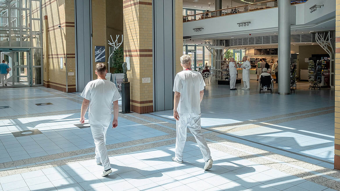 Zwei Klinikmitarbeiter gehen durch lichtdurchflutetes Krankenhausatrium mit Glasdach.