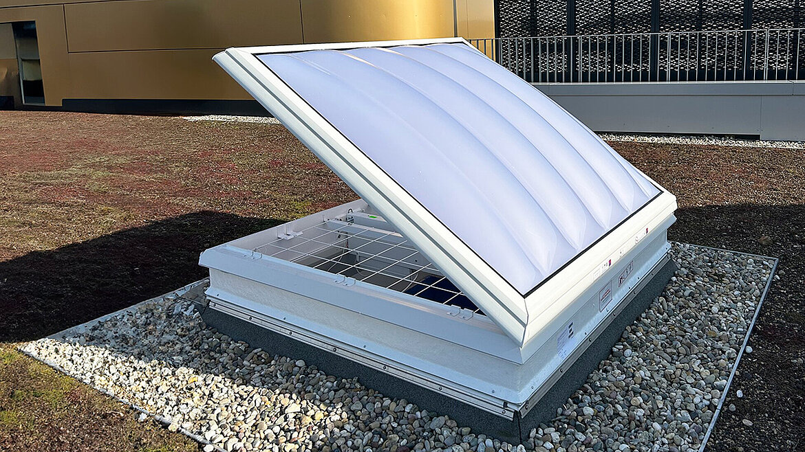 Geöffnete Lichtkuppel mit weißer Polycarbonat-Kuppel auf Flachdach mit Kiesumrandung und Gründach