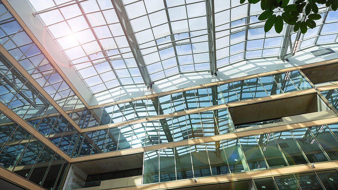 Modernes Gebäude-Atrium mit großem Glasdach, mehrstöckige Glasfassaden, Sonnenlicht und grüne Pflanze