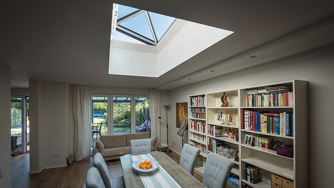 Modernes Wohnzimmer mit Oberlicht, Esstisch mit Stühlen, weißem Bücherregal und Panoramafenstern zum Garten
