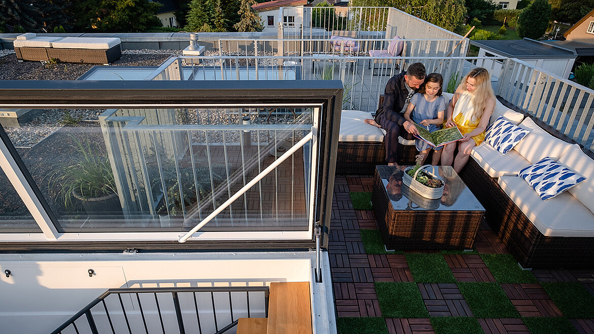Familie sitzt auf einer modernen Dachterrasse mit Lounge-Möbeln, umgeben von Pflanzen, mit Zugang über einen Flachdachausstieg.