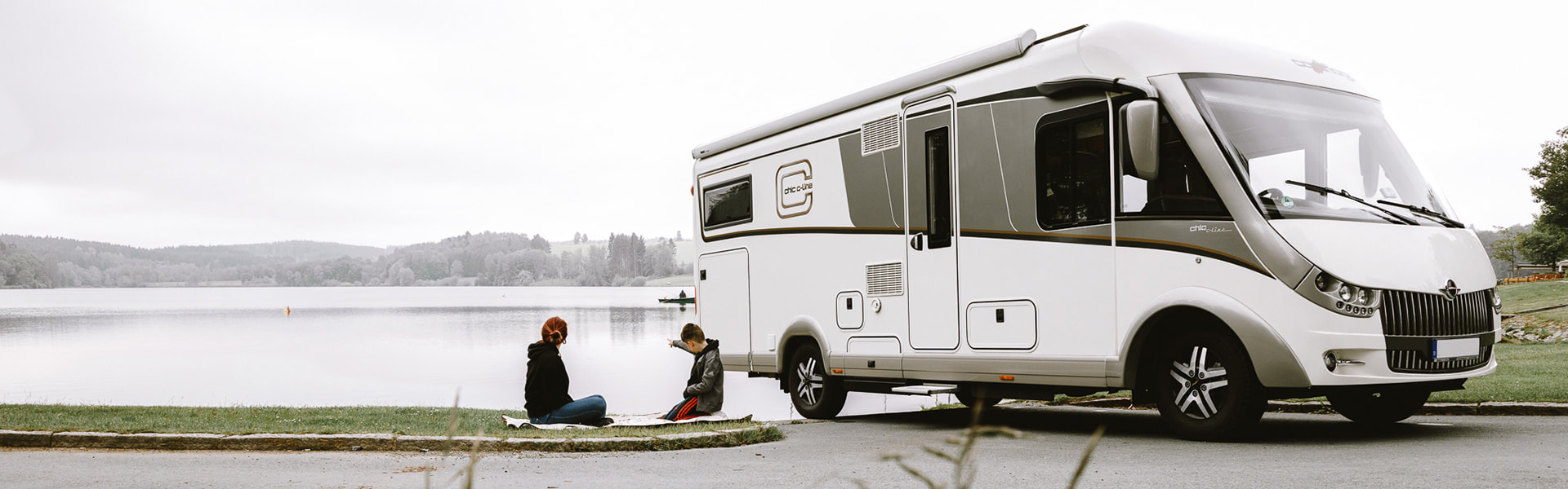 Wohnmobil am Seeufer mit zwei Personen, die auf der Wiese sitzen.